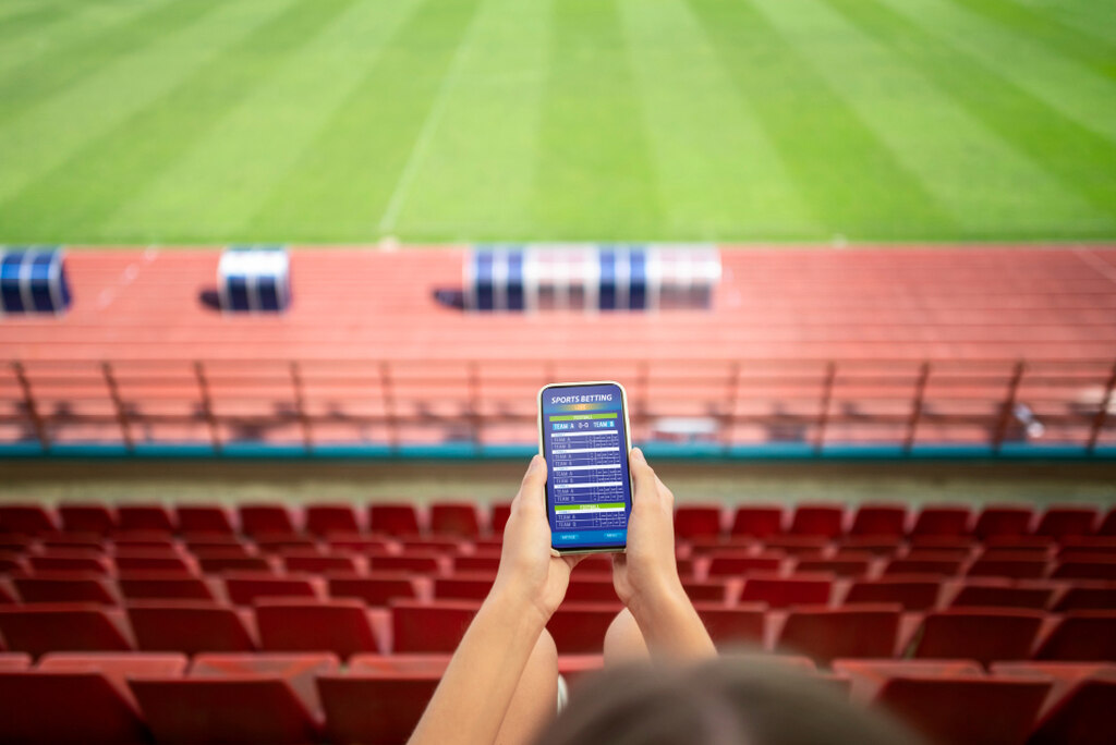 Pessoal sentada na arquibancada de um estádio, olhando para o gramado, segurando um celular com ambas as mãos.