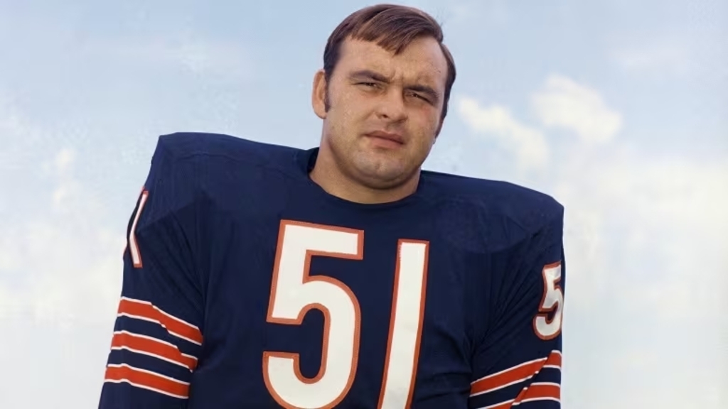 Ex-jogador de futebol americano, Dick Butkus, vestindo uniforme azul-escuro com detalhes vermelhos e brancos.