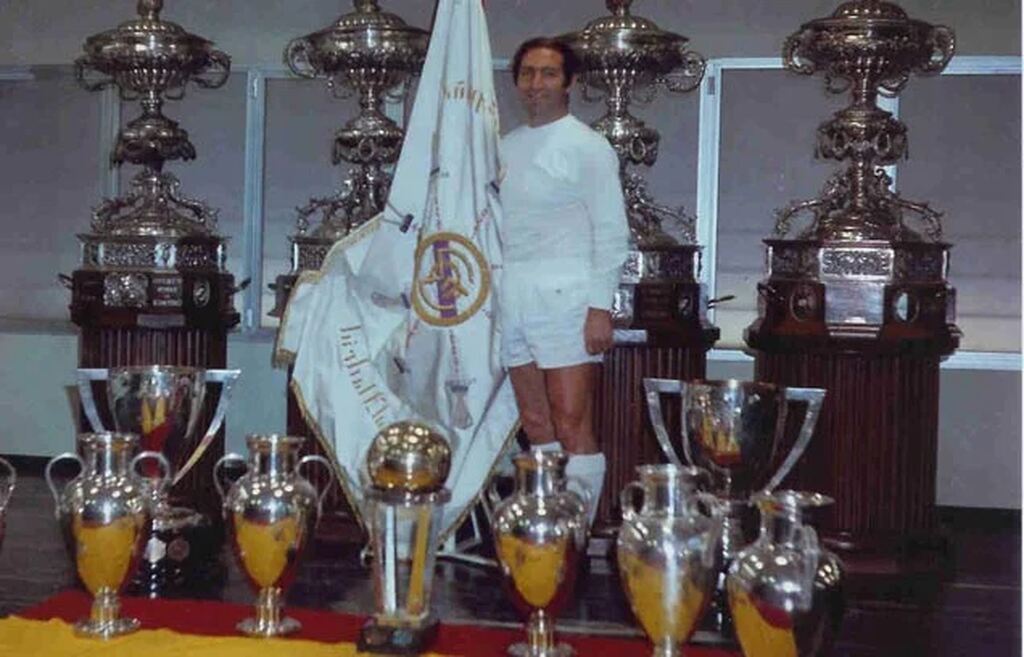 Francisco Gento, vestido uniforme branco do Real Madrid em galeria de troféus com bandeira do clube.