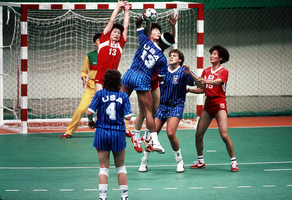 Jogadora de handebol, de uniforme azul dos EUA, realizando arremesso enquanto adversário de vermelho tenta bloquear.