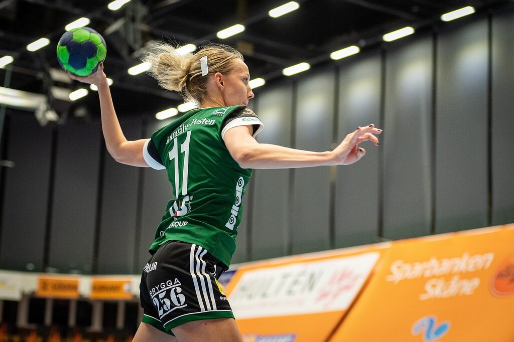 Jogadora de handebol vestindo uniforme verde, arremessando a bola contra o gol.