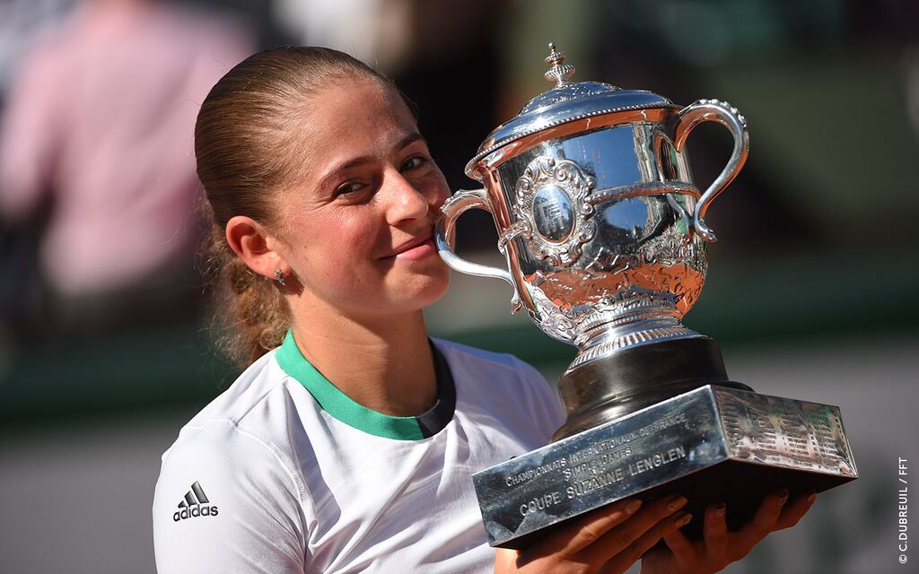 Jogadora de tênis Jelena Ostapenko, vestindo uniforme branco, erguendo taça prateada em comemoração.