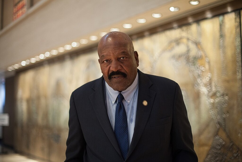 Ex-jogador de futebol americano, Jim Brown, vestindo terno preto e grava azul durante evento.