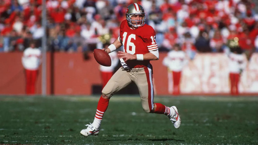 Ex-jogador de futebol americano, Joe Montana, vestindo uniforme vermelho e correndo com a bola durante partida.