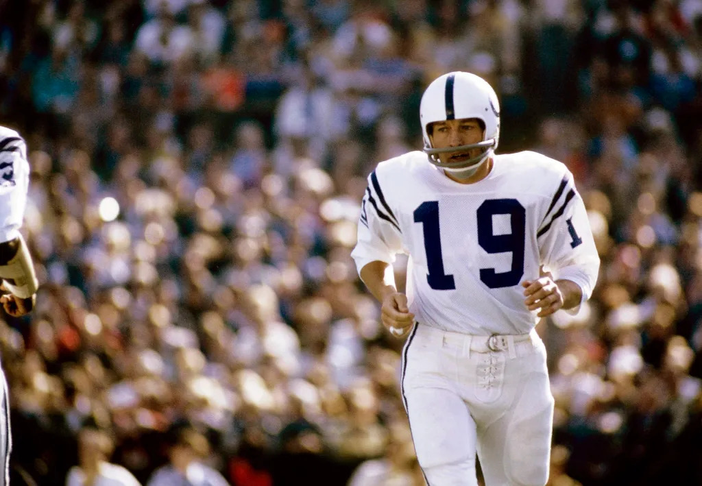 Ex-jogador de futebol americano, Johnny Unitas, vestindo uniforme branco com detalhes azuis, correndo durante partida.