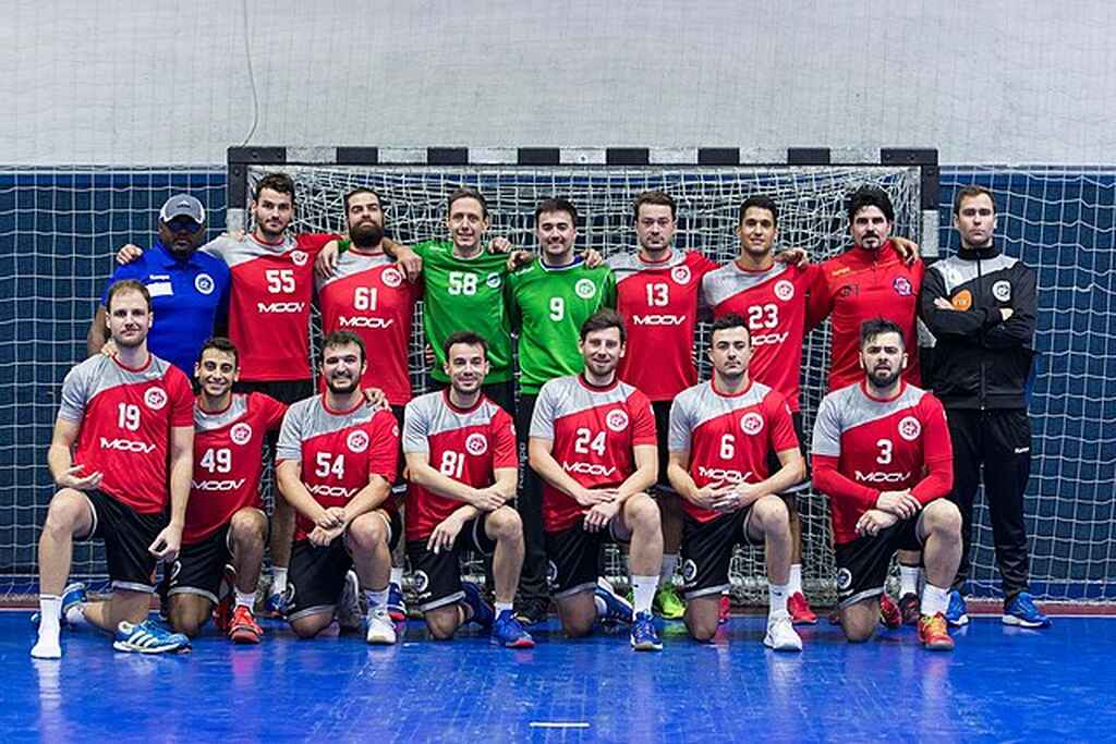 Equipe de handebol vestindo uniforme cinza e vermelho, abraçados em frente ao gol antes do início da partida.