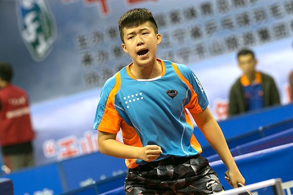 Jogador de tênis de mesa Wang Chuqin, de uniforme azul com detalhes laranjas, durante partida profissional.