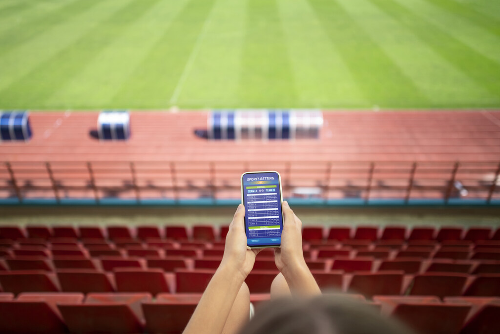 Pessoa segurando um celular com aplicativo de apostas esportivas aberto, em arquibancada de estádio de futebol.