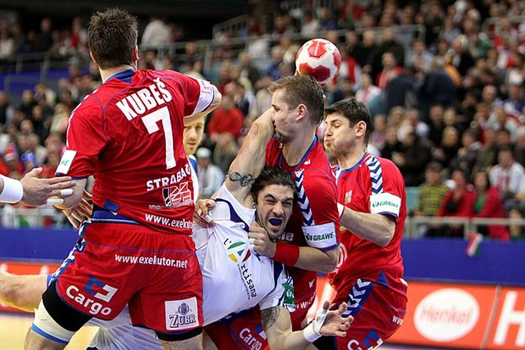 Jogador de handebol de uniforme branco, sendo marcado por três jogadores de uniforme vermelho durante arremesso.