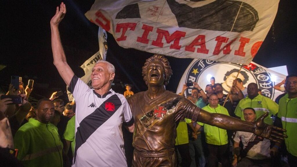 Roberto Dinamite, vestindo camisa branca com listra preta do Vasco, abraçado com sua estátua.