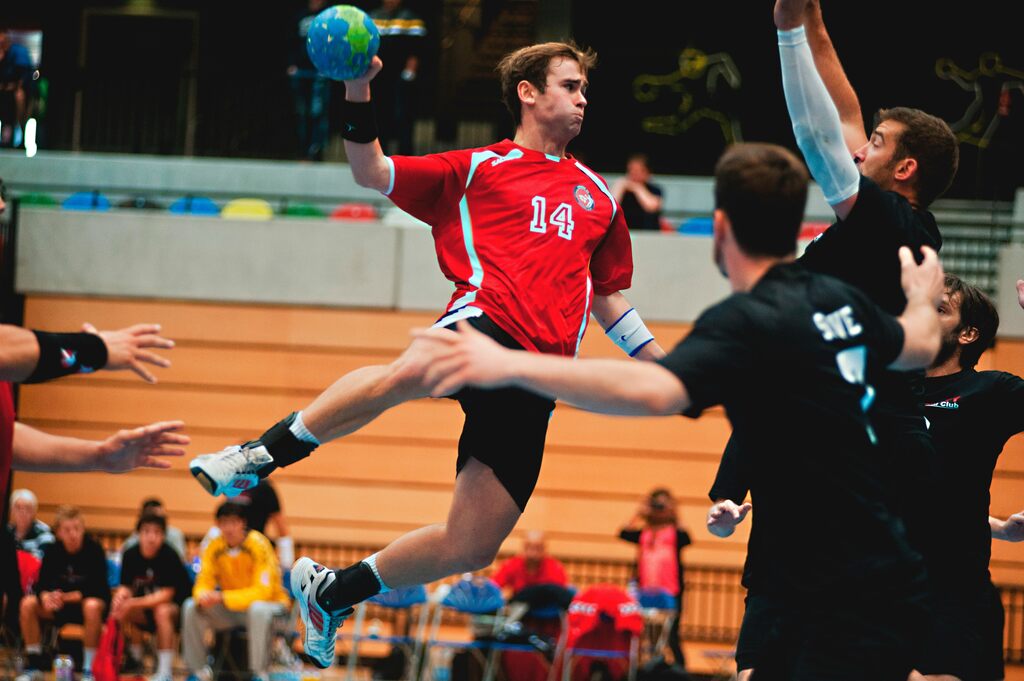 Jogador de handebol vestindo uniforme vermelho com detalhes brancos, realizando um arremesso com a mão esquerda.