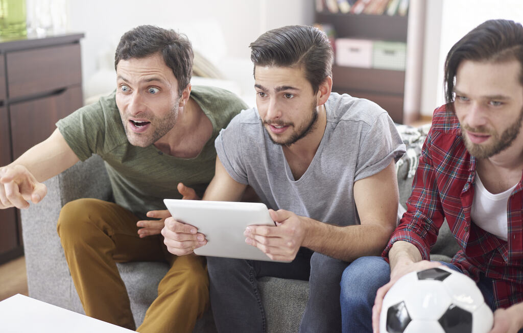 3 homens assistindo a partida de futebol enquanto seguram um tablet e uma bola de futebol.