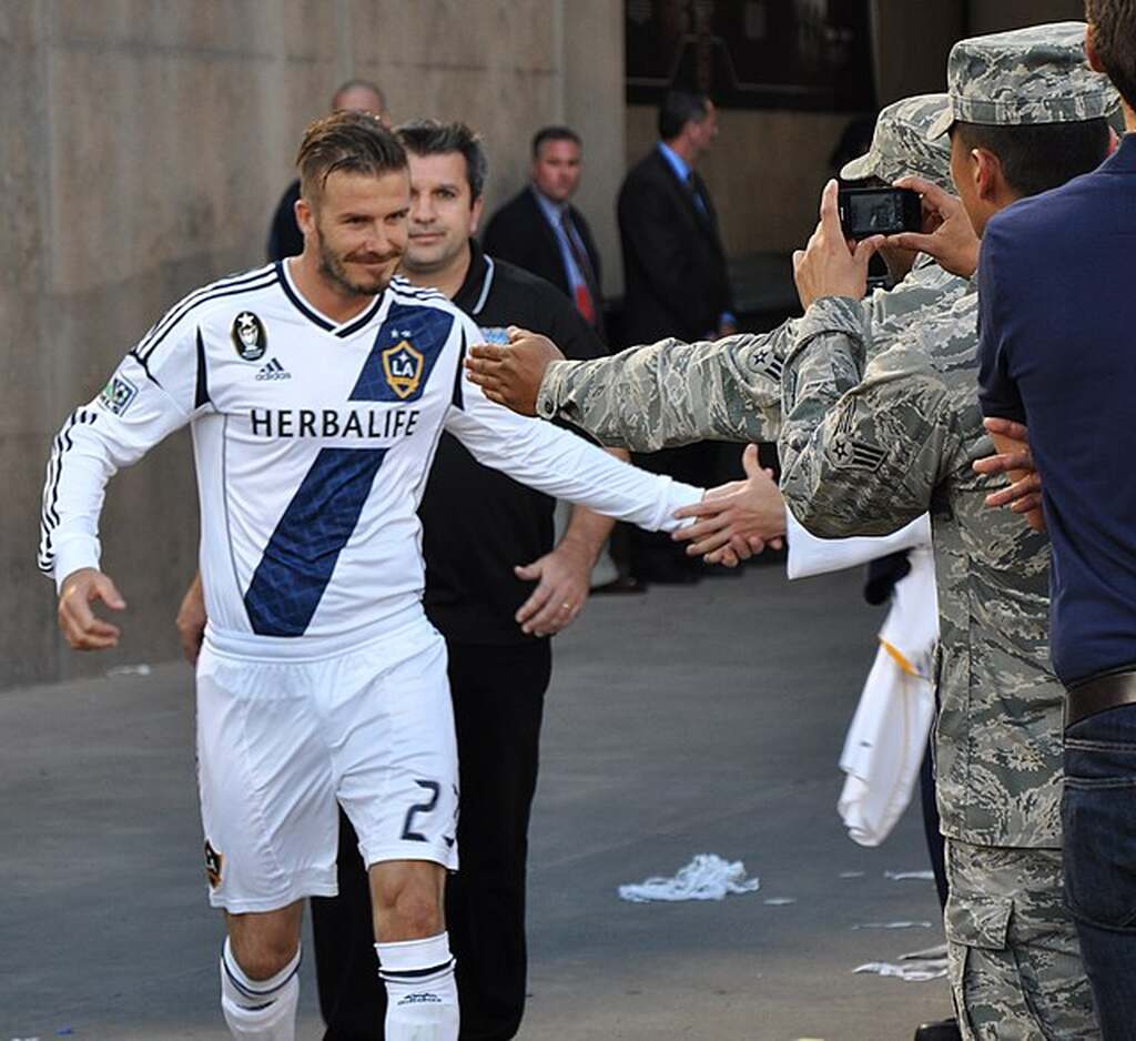 Ex-jogador de futebol, David Beckham, vestindo uniforme branco com detalhes azuis, entrando em campo na MLS.