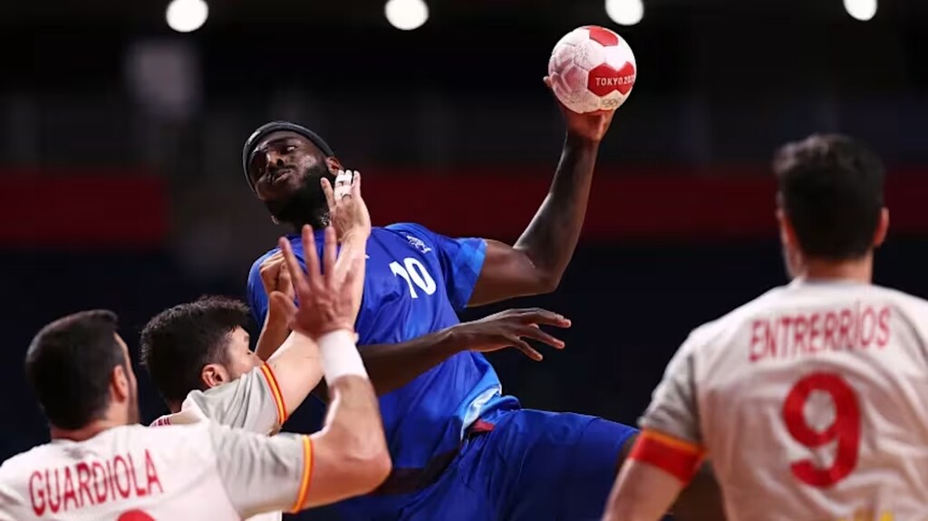 Jogador da seleção francesa de handebol, vestindo uniforme azul, arremessando bola durante partida contra a Espanha.