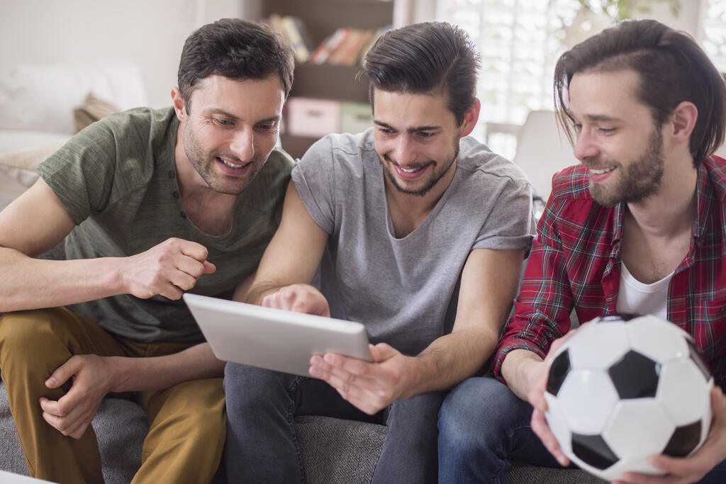 3 homens sentados em um sofá, um deles segurando uma bola, enquanto fazem apostas esportivas em um tablet.