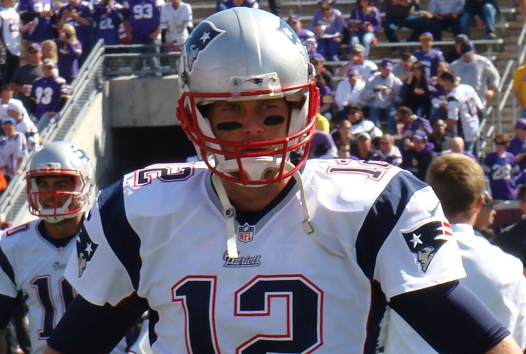 Jogador de futebol americano, Tom Brady, vestindo uniforme branco com detalhes pretos e usando capacete em sua entrada ao estádio.