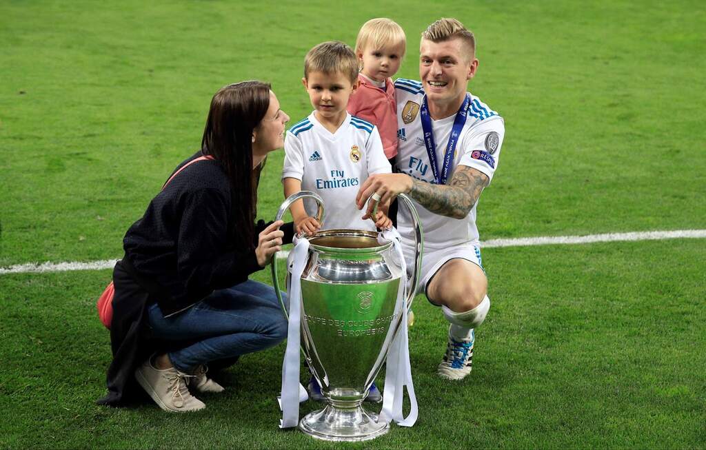 Jogador de futebol Toni Kroos, vestindo uniforme do Real Madrid com seus filhos e esposa perto da taça da Champions.