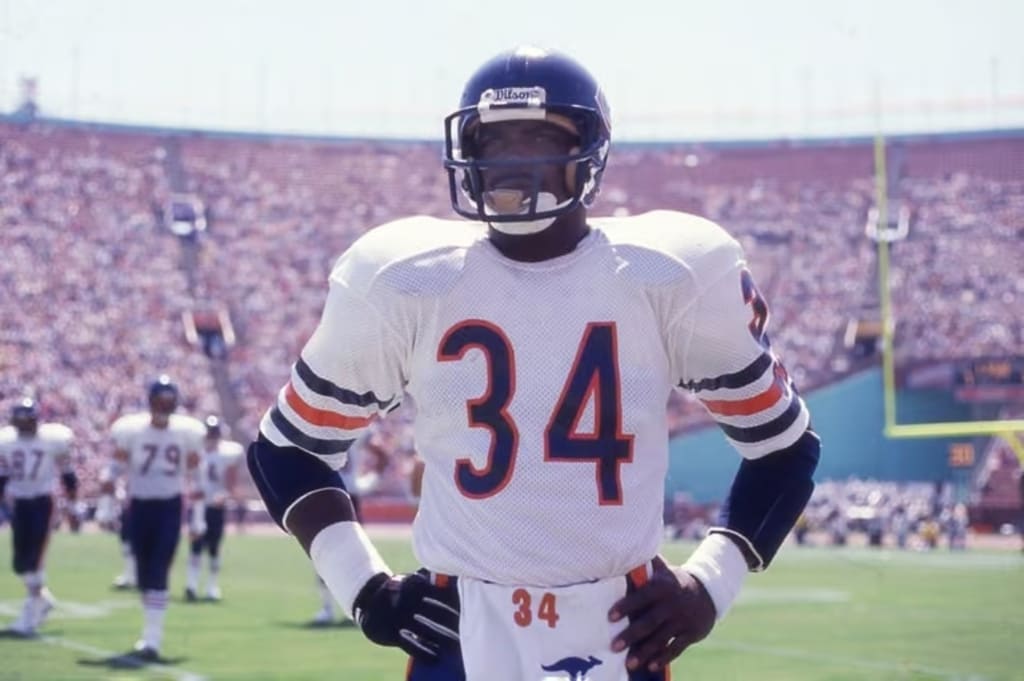 Ex-jogador de futebol americano, Walter Payton, vestindo uniforme branco com detalhes azuis e vermelhos durante partida.