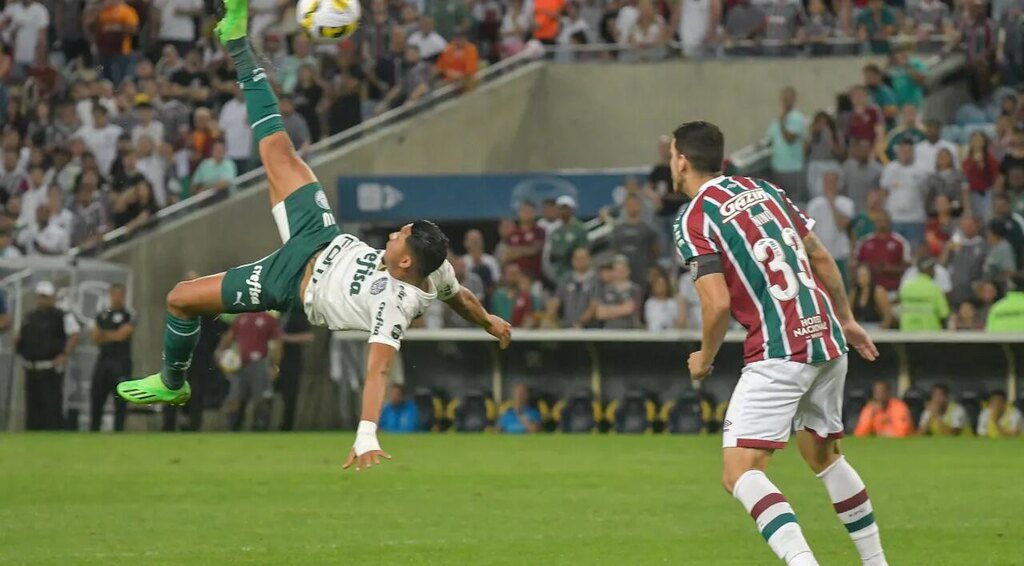 Jogador de futebol do Palmeiras, Rony, usando uniforme branco e verde, realizando bicicleta durante partida.