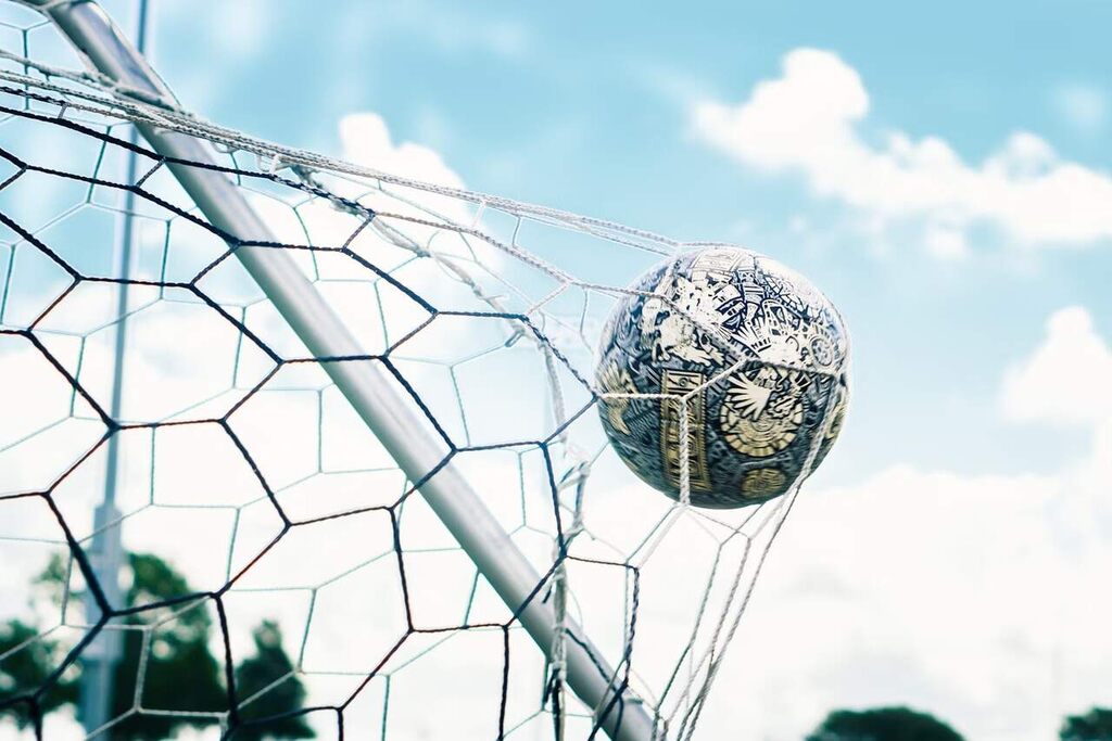 Bola de futebol dentro do gol, empurrando a rede mais para o fundo durante dia de sol.