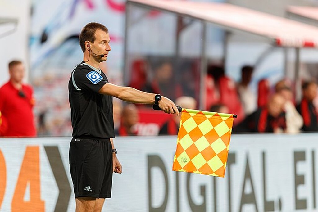 Bandeirinha de futebol, segurando a bandeira amarela com listras laranjas e vestindo uniforme preto, durante partida de futebol.