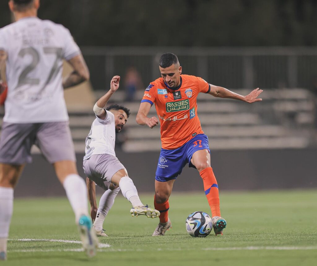 Jogador brasileiro Ricardo Ryller, vestindo uniforme laranja e realizando drible durante jogo pelo Al-Fayha.