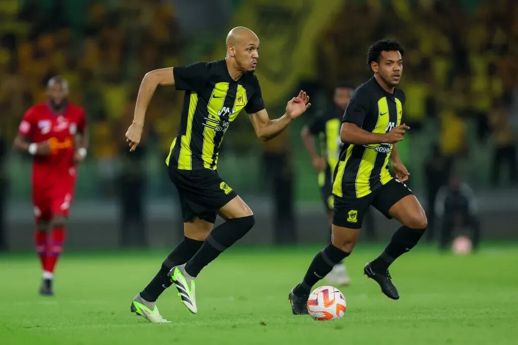 Fabinho e Romarinho vestindo uniforme preto e amarelo do Al-Ittihad durante parida de futebol.