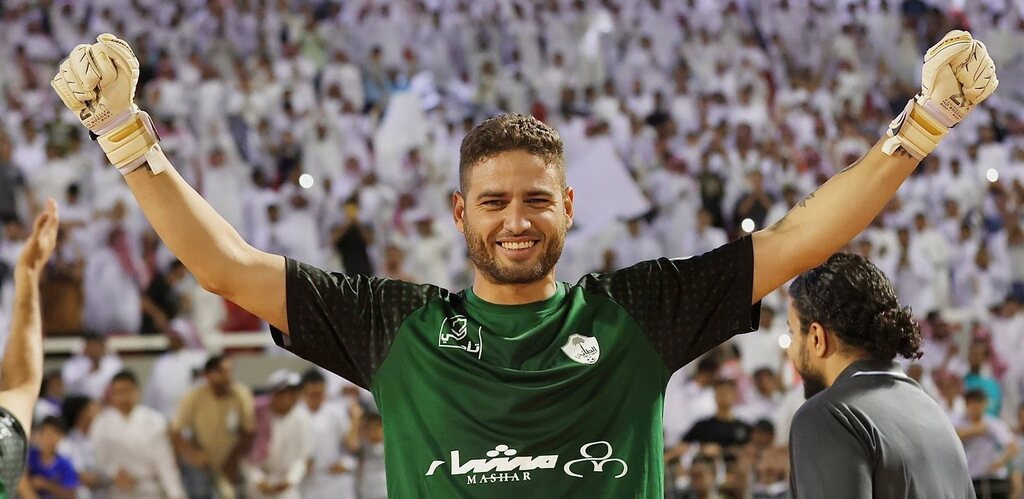 Goleiro Victor Braga vestindo uniforme verde do Al-Tai FC, com os braços para o alto comemorando algo.