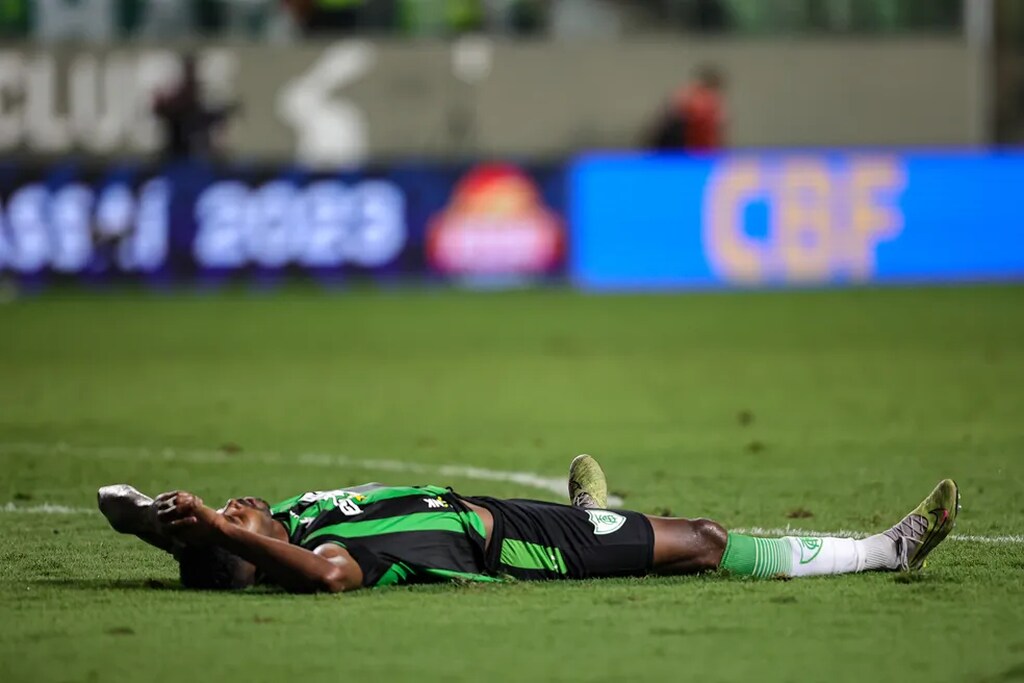 Jogador do América-MG, vestindo uniforme verde com detalhes preto, deitado no gramado durante partida.