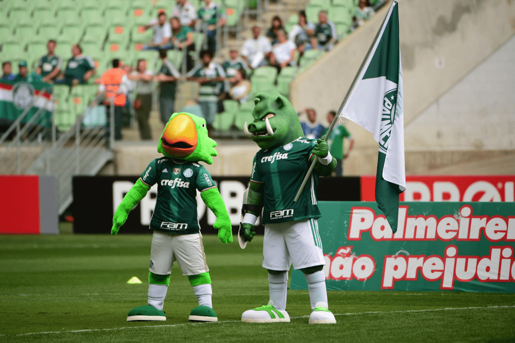 Mascotes do Palmeiras segurando a bandeira do time no gramado durante partida de futebol.