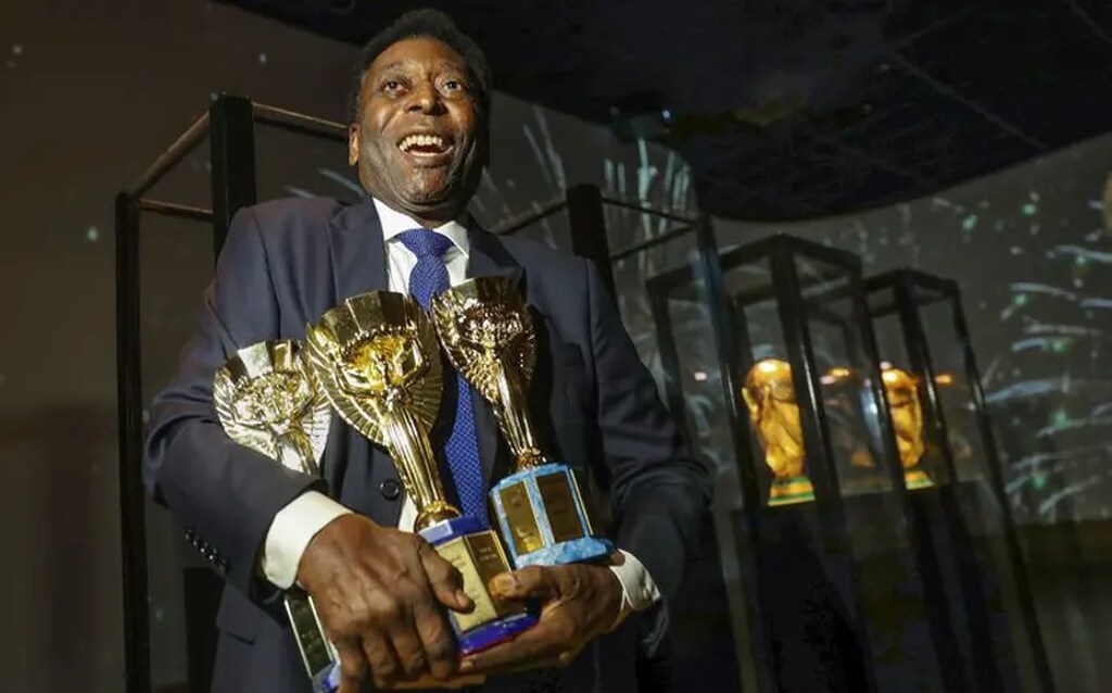 Rei do futebol, Pelé, vestindo roupa social enquanto segura as 3 taças da Copa do Mundo de quando foi campeão.