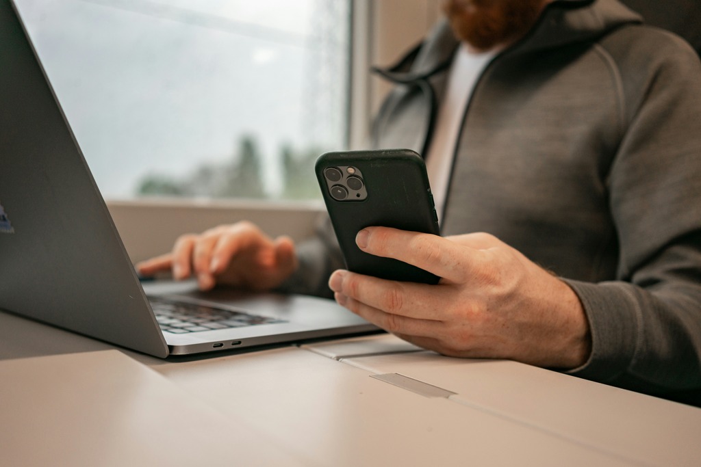 Homem de casaco preto com blusa branca usando um notebook e segurando um celular com a mão esquerda.