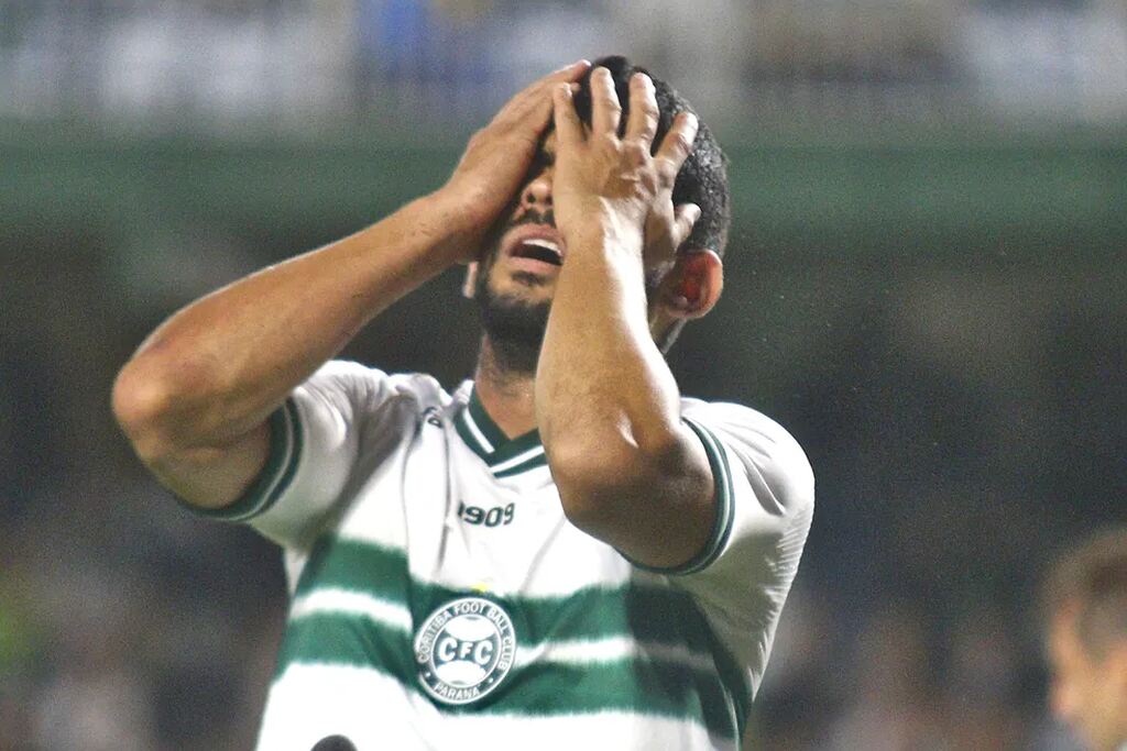 Jogador do Coritiba, vestindo camisa branca com detalhes verdes, levando a mão ao rosto frustrado.