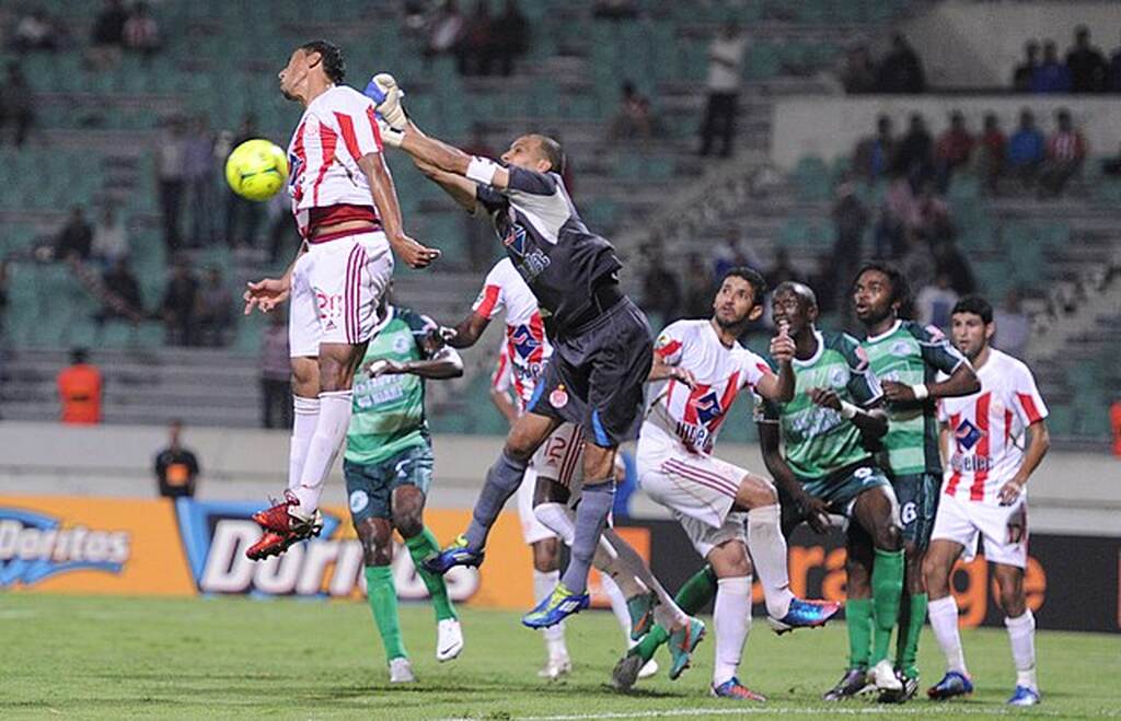 Goleiro disputando bola no ar com jogador adversário durante cobrança de bola parada.