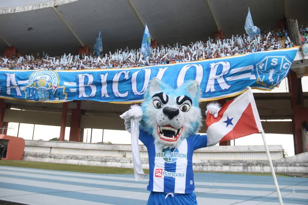 Mascote do Paysandu, com uniforme azul e branco, em frente a torcida do time durante partida.