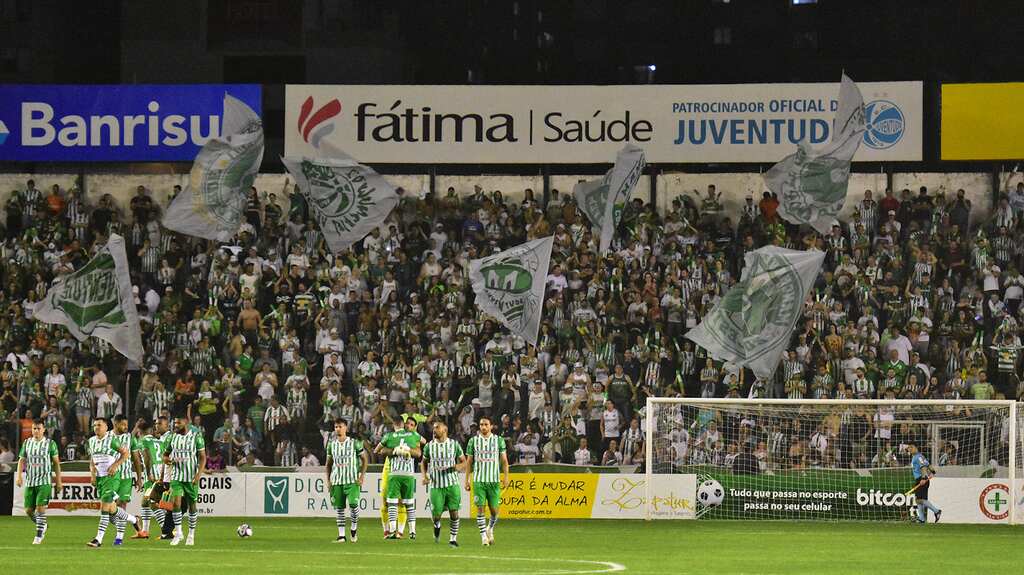 Arquibancada do estádio do Juventude lotado, com torcida vestindo verde e branco erguendo bandeiras durante o jogo.