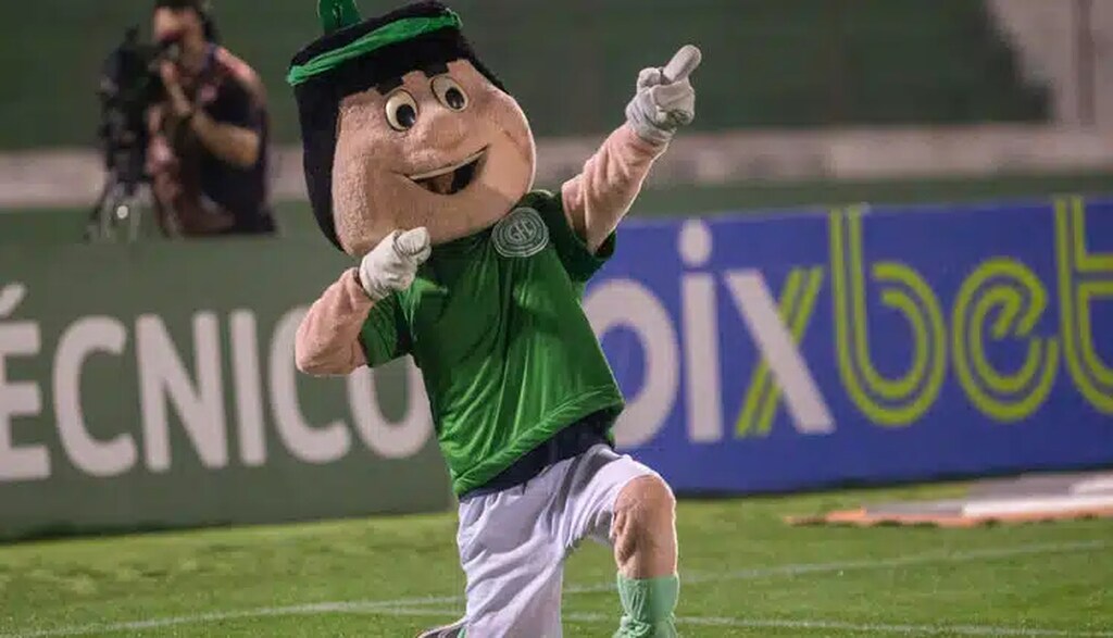 Mascote do Guarani vestindo camisa do clube e calção branco no gramado de futebol.