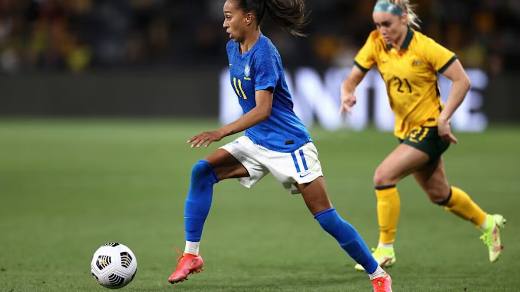 Jogadora da seleçõ de futebol feminino do Brasil, conduzindo a bola enquanto é marcada por jogadora adversária.