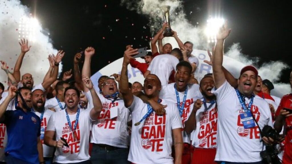 Imagem dos jogadores do Náutico sendo campeões da Série C em 2019, com os jogadores sorrindo e com as mãos para o alto comemorando a vitória. Um deles ergue a taça.