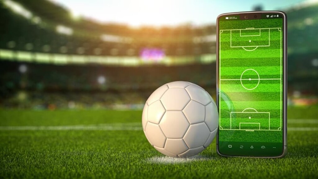 Uma bola de futebol ao lado de um celular com uma imagem de um campo de futebol, e um estádio ao fundo