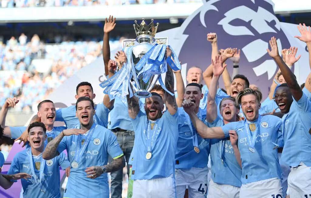 Jogadores do Manchester City comemorando a vitória na Premier League, pulando sorridentes e um deles ergue a taça do campeonato